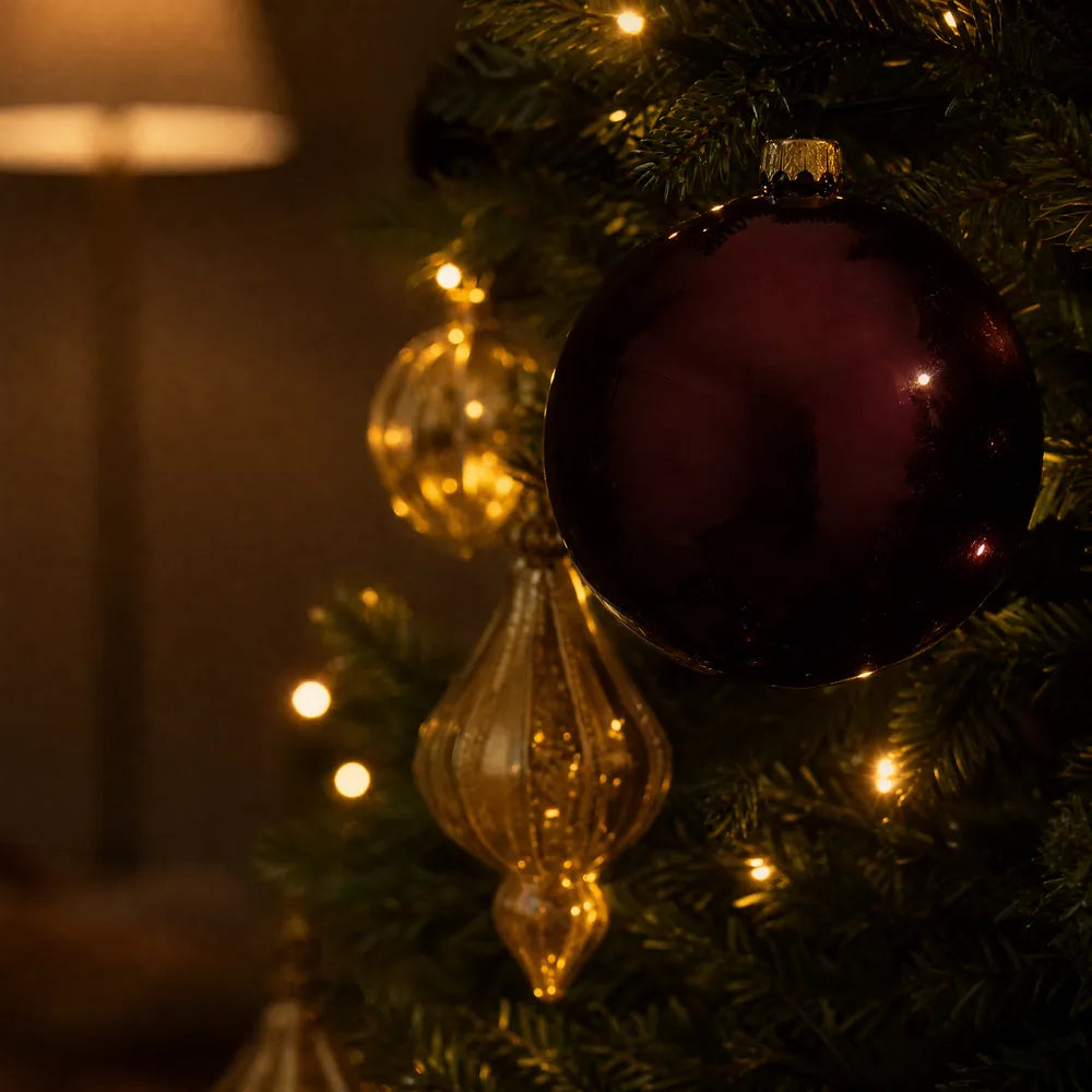 A burgundy glass ornament close up on a branch, a gold ornament and small warm lights softening away behind it.