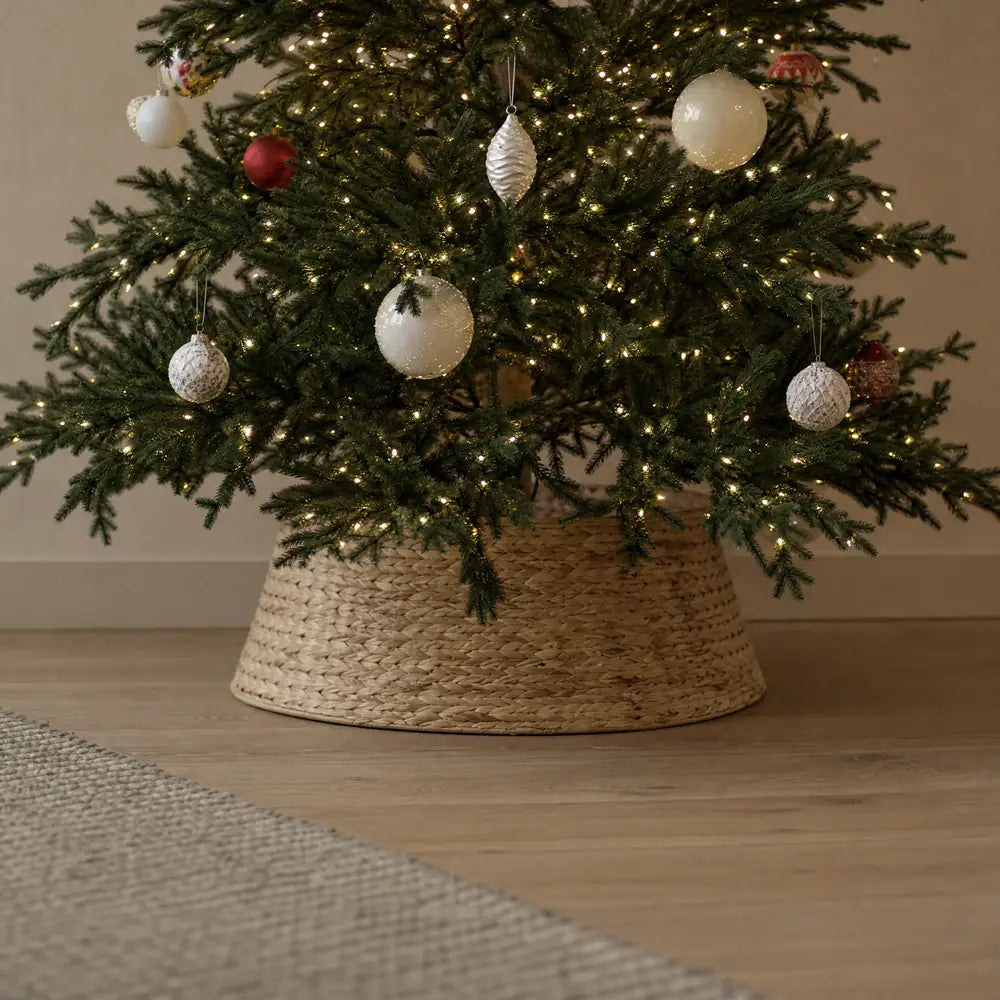 A woven collar around the foot of the tree on pale oak flooring, the lit lower branches above it carrying white, frosted and red ornaments.