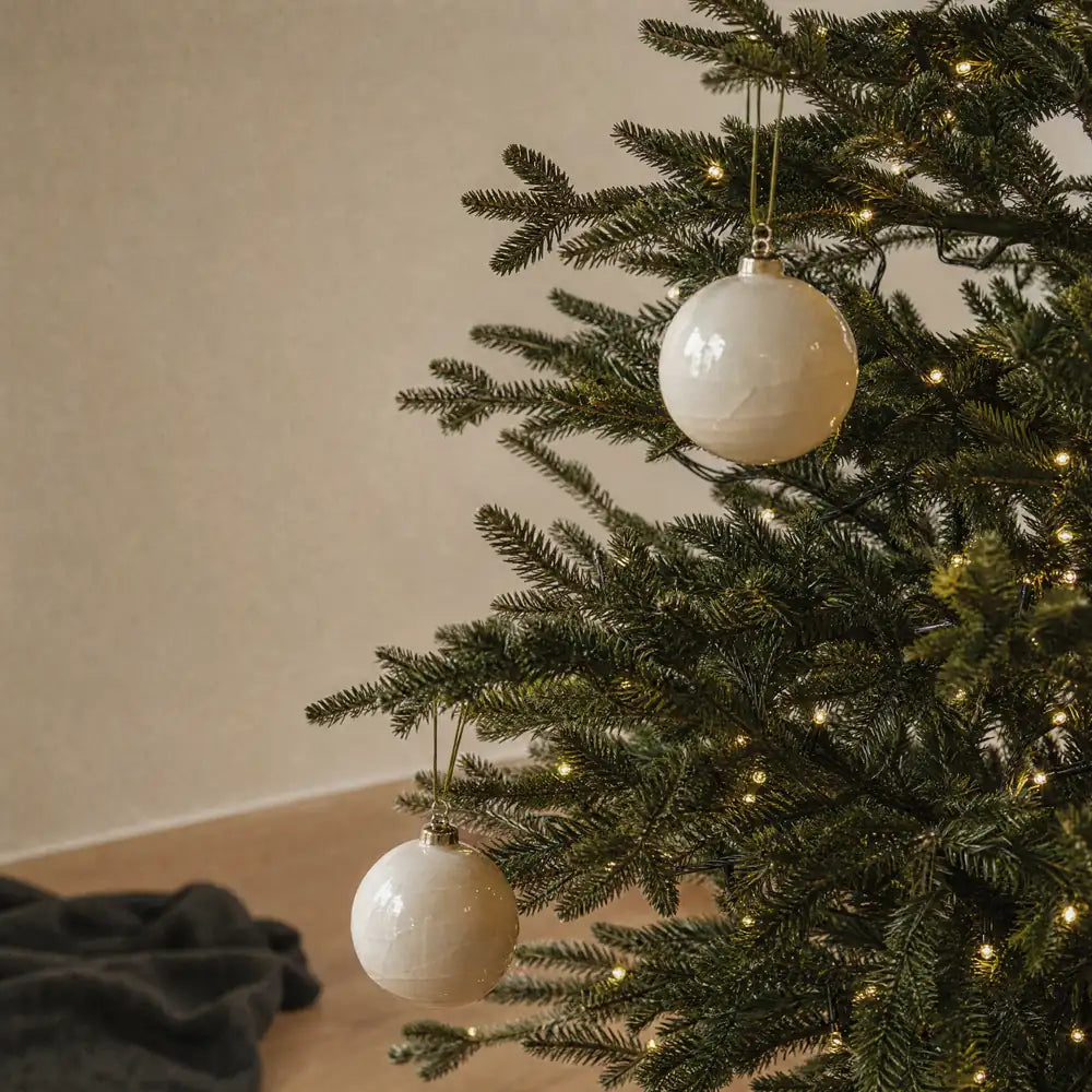 Two pale glazed ornaments hanging on open fir branches against a bare oatmeal wall, a charcoal throw out of focus at the lower left.